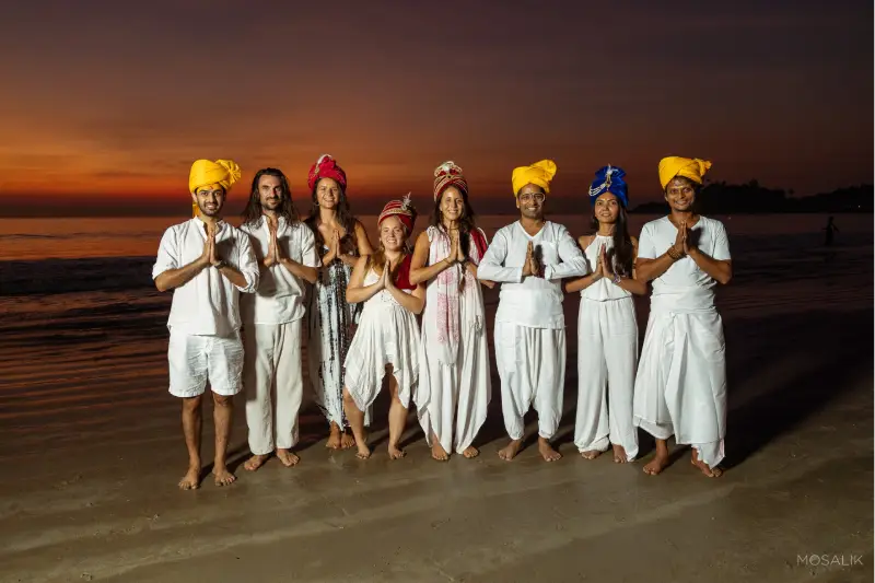 A group of people participating in a 200 Hr Ashtanga & Vinyasa TTC training pose for a photo on the beach.